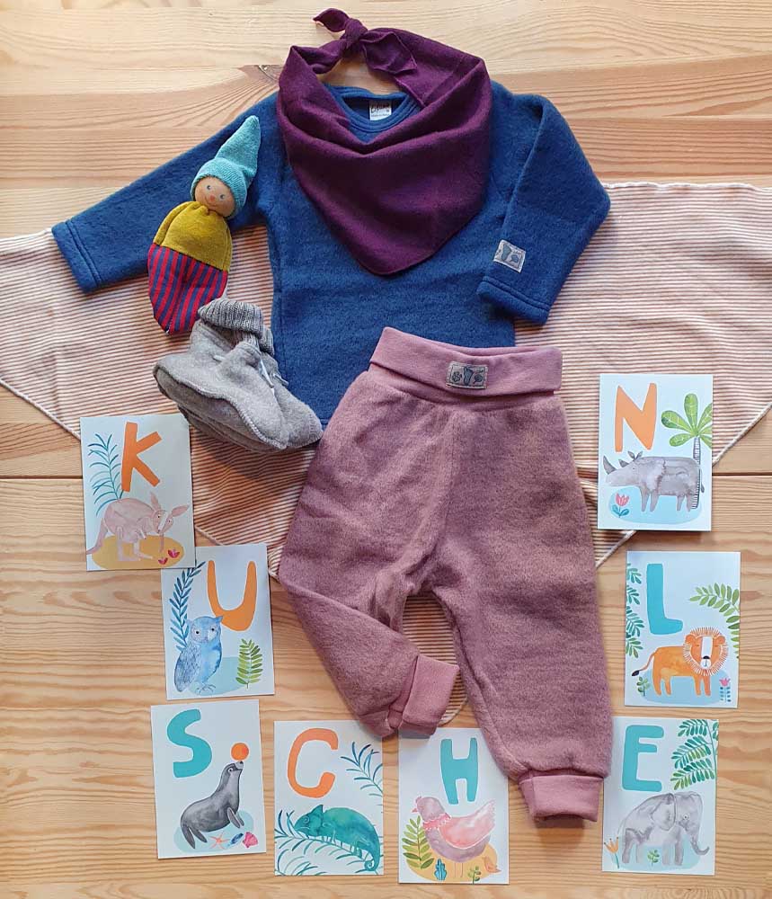 Lilalo Kleidung für ein Baby: Pullover aus Wollfleece in blau kombiniert zu einer Pumphose aus Wollfleece in mauve. Als Accessoire das Dreieckstuch aus Wolle/Seide in zwei verschiedenen Größen. Dazu passen die Wollschuhe von Joha, eine Rassel als Puppe von Nanchen Natur und ABC-Karten von Frau Ottilie, die das Wort kuscheln ergeben.