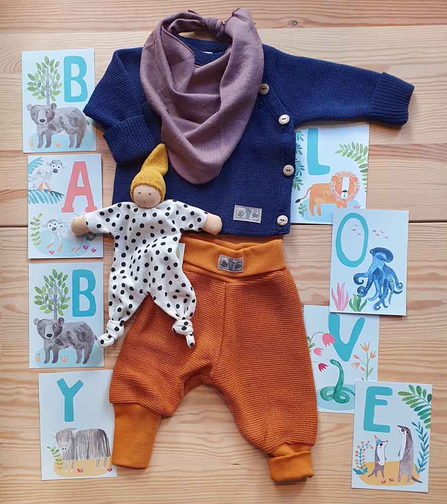 Flatlay mit einem Outfit von Lilano. Blauer Pullover und braune Pumphose aus Wolle mit einem Dreieckstuch aus Wolle/Seide in mauve. Passend dazu eine Kuschelpuppe von Nanchen Natur und ABC-Karten von Frau Ottilie, die die Worte Baby Love ergeben.