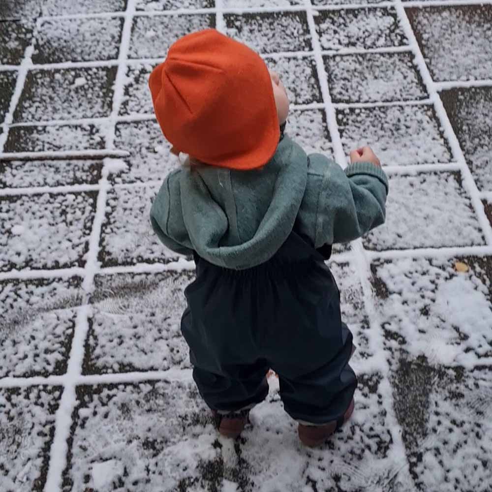 Kind steht in einem Wollwalkanzug von Engel Natur, einer Mütze aus Wolle von Disana und einer Regenhose im Schnee