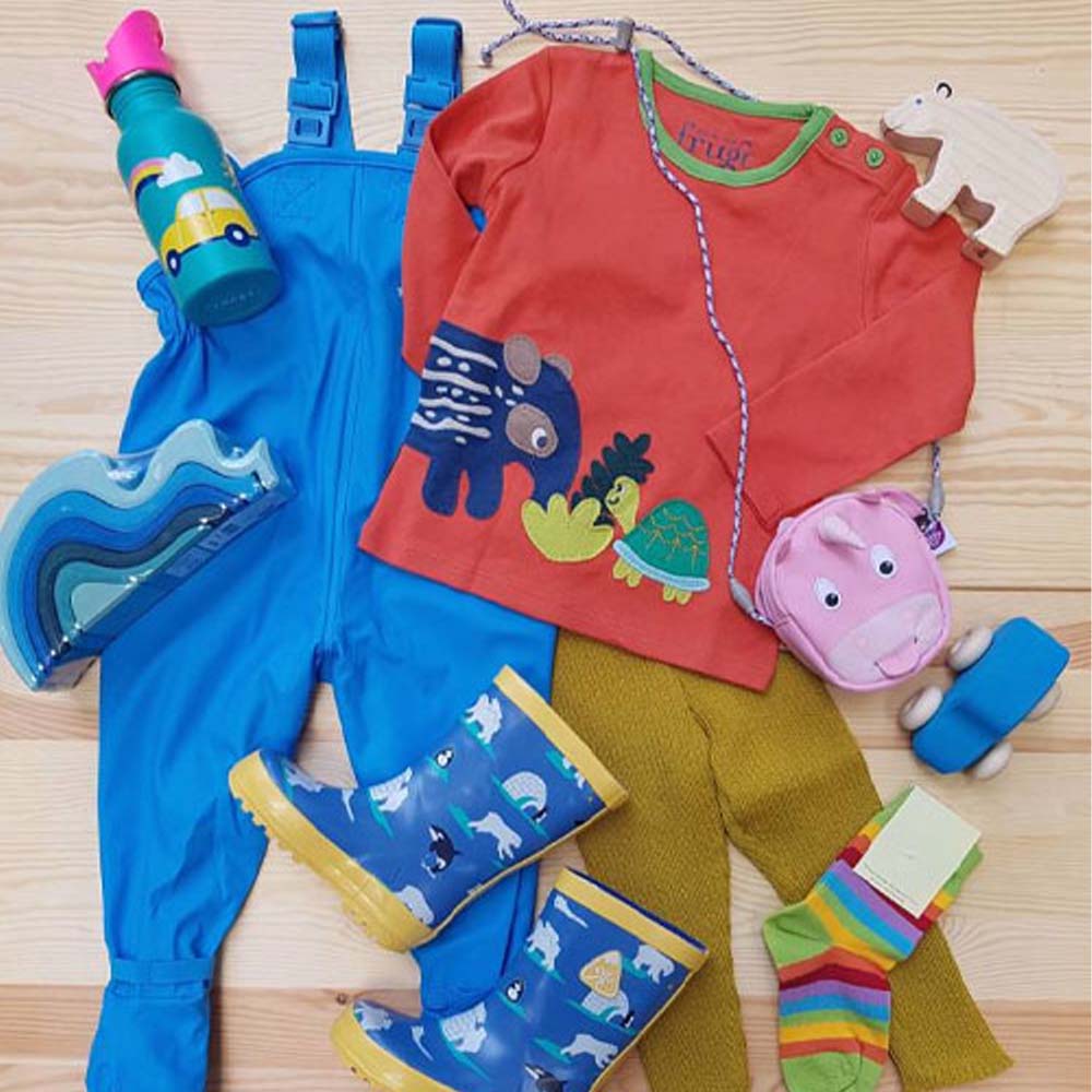 Flatlay mit Kinderkleidung: Matschhose in blau, Gummistiefel mit EIsbären-Motiv, Langarmshirt von Frugi mit Tieren und einer gelben Wollhose von Disana.