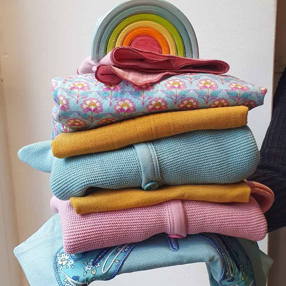 Stapel mit festlicher, nachhaltiger und fair produzierter Kinderkleidung von Enfant Terrible. Oben drauf thront ein kleiner Regenbogen von Grimms Holzspielzeug.