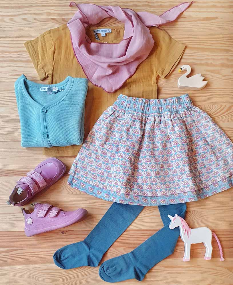 Festliches Outfit mit Kleidung: Flatlay mit einem Wenderock, T-Shirt mit Blumen-Stickerei, einer Strumpfhose & Cardigan in blau sowie dazu passenden pinken Affenzahn Schuhen & Musselintuch. Dazu drapiert sind ein Einhorn und Schwan aus Holz von Holztiger.